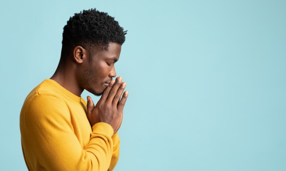 Side view of praying african american young man in yellow, holding palms by his lips and whispering pray, asking for grace over blue studio background, panorama wuth copy space
